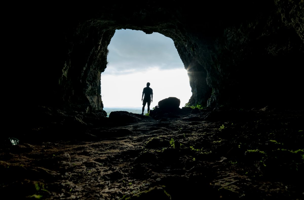 From inside a dark cave, the camera looks out at the cave's circular exit at grey cloudy sky and the outline of the sea. The figure of a person standing at the cave's entrance is in silhouette.